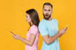 Side view of concerned young couple two friends guy girl in blue pink t-shirts isolated on yellow background. People lifestyle concept. Standing back to back, using mobile phones, typing sms messages.