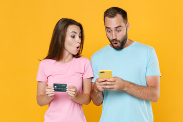 Wall Mural - Shocked young couple two friends guy girl in blue pink t-shirts isolated on yellow background studio portrait. People lifestyle concept. Mock up copy space. Using mobile phone, hold credit bank card.