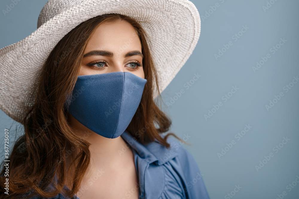 Woman wearing stylish protective face mask, posing on blue background. Trendy Fashion accessory during quarantine of coronavirus pandemic. Close up studio portrait. Copy, empty space for text - obrazy, fototapety, plakaty 