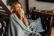 Sick woman covered with blanket sneezing in napkin while sitting on sofa