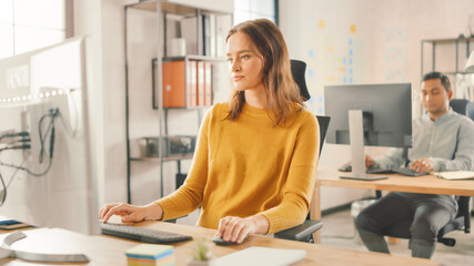 Beautiful and Smart Red Haired Female Specialist Sitting at Her Desk Works on a Desktop Computer. In the Background Modern Bright Office with Diverse Group of Professionals Working for Growing Startup