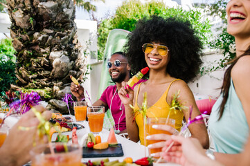 Black young woman smiling at the camera celebrating with friends. Diverse people hanging out toasting and drinking cocktails outdoor.