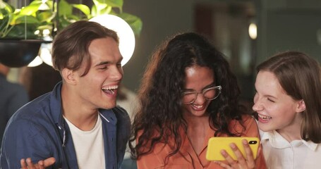 Close up view of young people using smartphone and laughing while enjoying social media app. Group of diverse students having fun together while looking at phone screen. Indoors.