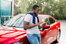 Cheerful Casual Hipster Black Man Using Smart Phone And Writing Text Messages, Standing Near His Modern Red Car Outdoors
