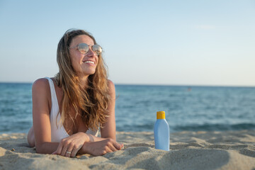 An young smiling woman in white bikini and sunglasses is lying on sandy beach of a seaside with sunscreen or sun tanning lotion anonime bottle behind to take care of her skin during holidays vacation.