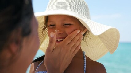 Wall Mural - Close up of young mother is applying protective sunscreen or sunblock lotion on her little happy smiling daughter's face to take care of skin on a seaside beach during family holidays vacation.