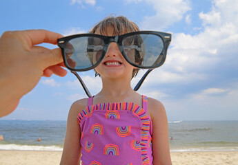 Someone holding sunglasses, and little girl on the beach, smiling and looking through it.