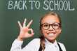 © LIGHTFIELD STUDIOS - selective focus of excited schoolgirl in eyeglasses showing thumb up near chalkboard with back to school text
