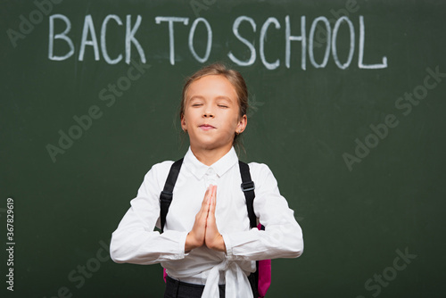 cute schoolgirl with closed eyes holding praying hands near back to school lettering on chalkboard