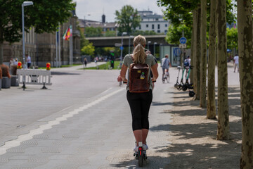 Wall Mural - One girl ride E-scooters, trendy urban transportation with Eco friendly sharing  mobility concept, on bicycle lane at promenade riverside of Rhine River in Düsseldorf, Germany.