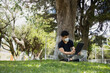 © Fernanda - Hispanic young man sitting reading in the park with mask - young man studying with an electronic tablet in a natural park - social distancing - general shot