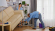 © DC Studio - Young woman helping husband to clean the dust under sofa.