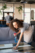 © pressmaster - Content curly-haired girl in apron wiping coffee table with cloth while disinfecting surfaces in cafe