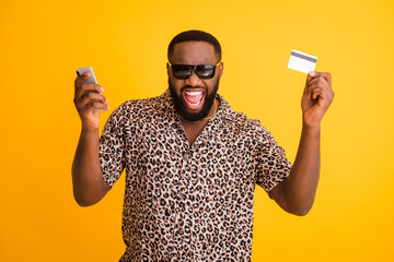 Photo of crazy funny dark skin guy hold telephone make online payment purchase transfer use credit card wear sun glasses golden necklace leopard shirt isolated bright yellow background