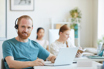 Wall Mural - Portrait of bearded businessman in headphones smiling at camera while working at the table with laptop among his colleagues at office