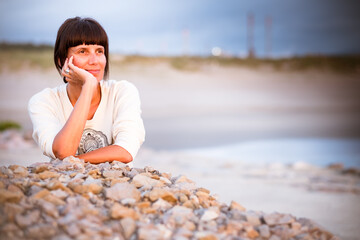 Mujer de mediana edad al atardecer observa el horizonte con la playa de fondo y desenfocada