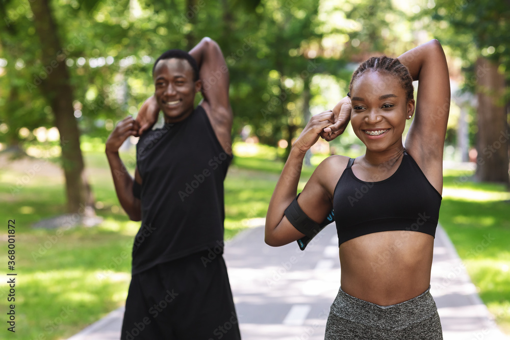 Sporty african couple exercising before jogging at park, stretching arm muscles outdoors - obrazy, fototapety, plakaty 