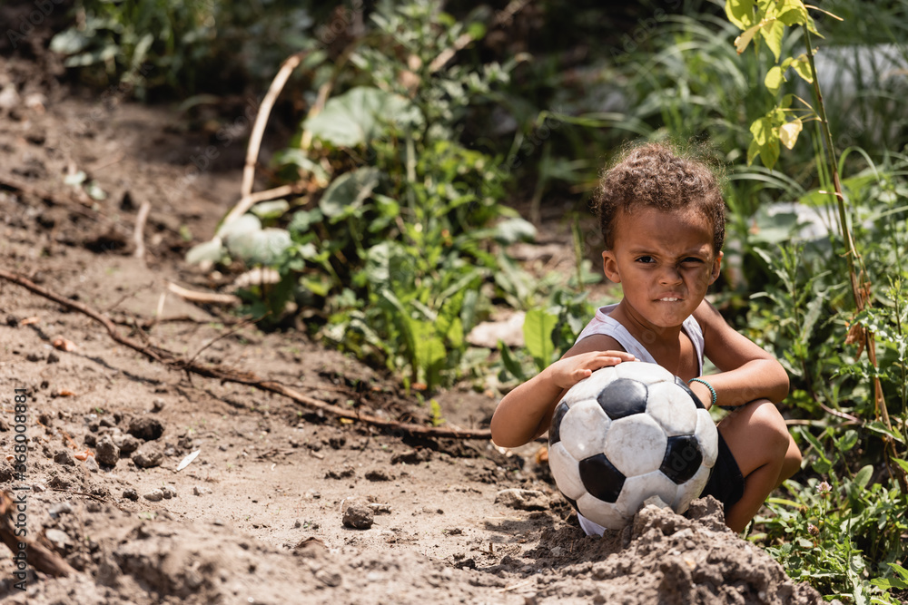 Poor african american boy holding football while sitting near plants on dirty road - obrazy, fototapety, plakaty 
