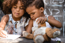 Selective Focus Of African American Kid Sticking Out Tongue While Writing Near Brother Playing With Dirty Teddy Bear