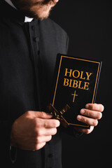 Canvas Print - cropped view of bearded priest holding cross and holy bible isolated on black