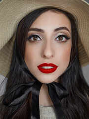 Wall Mural - Close up headshot portrait of young natural looking casual beautiful brunette woman with trendy makeup and vibrant red lips in straw hat liten by natural light domestic room background