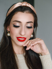 Wall Mural - Close up headshot portrait of young natural looking casual beautiful brunette woman with trendy makeup and vibrant red lips in hair hoop and pearl earrings domestic room background