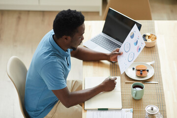 African young businessman sitting at the table in front of laptop and examining documents during his breakfast at home