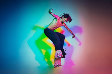 Skateboarder doing a trick isolated on studio background in colorful neon light. Young man shirtless riding and skateboarding in motion. Concept of leisure activity, sport, extreme, hobby and motion.