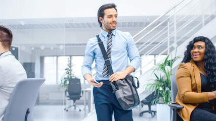 Young Happy Businessman Walks Into Bright Modern Office, Holds Smartphone and Takes Place at His Desktop Computer. In the Background Diverse Team of Professional Businesspeople Working