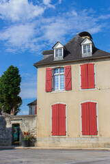 Old historical building with red window shutters and outdoor decor. Empty street in village, France. Facade of ancient house with tree in garden. Travel in France. Beautiful building on empty square.