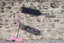 Signpost And Indication. Tilted Pink Road Sign With Black Board Arrow Showing Direction. Wall Background In Sunny Day. Signpost And Indication.
