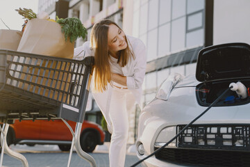 Charging electro car at the electric gas station. Woman standing by the car. Lady with foodstuff.