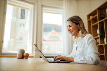 Wall Mural - Side view portrait of a cheerful young woman using laptop at home office.