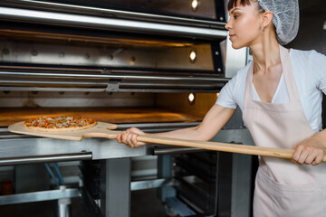 Wall Mural - Young caucasian woman baker is holding a wood peel with fresh pizza and put it in an oven at a baking manufacture factory.