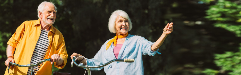 Panoramic shot of smiling elderly woman pointing with finger near husband and bikes in park