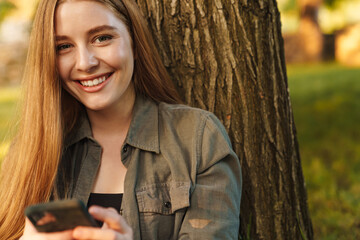 Young blonde woman using smartphone under tree in park