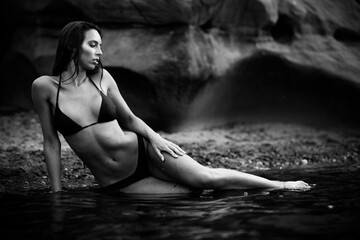 Wall Mural - Summertime recreation concept. Beautiful young sexy woman with fit trained slim body wearing black swimwear bikini lies on a beach. Fashion female model poses by the sea. Black and white monochrome.