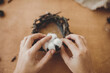 © sonyachny - Florist making rustic christmas candle holder. Hands holding cotton and making rustic festive decoration on wooden table with branches and pine cones. Holiday workshop