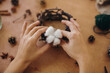 © sonyachny - Holiday workshop. Florist making rustic christmas candle holder. Hands holding cotton and making rustic festive decoration on wooden table with branches and pine cones.