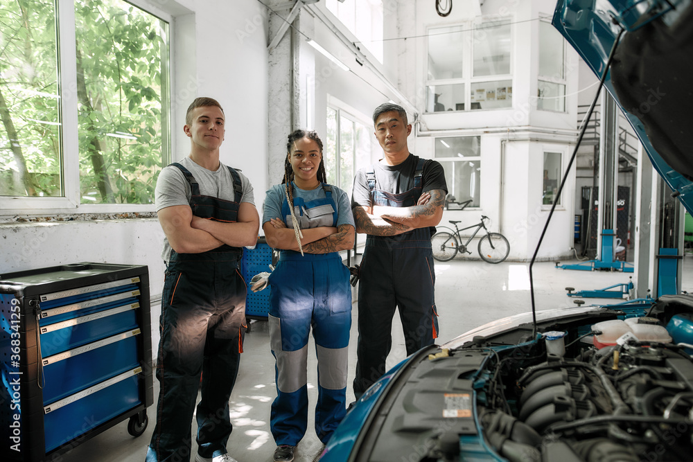 We value your car. Team of proud diverse mechanics in uniform, two men and a woman smiling at camera, while standing at auto repair shop