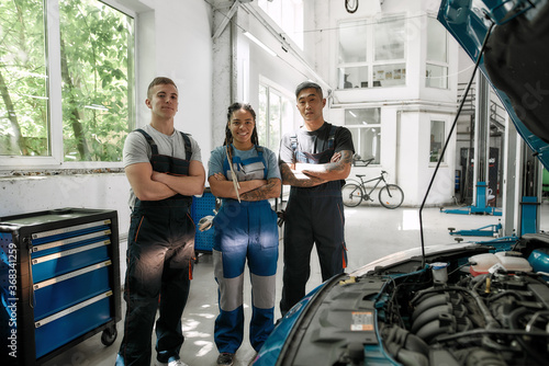We value your car. Team of proud diverse mechanics in uniform, two men and a woman smiling at camera, while standing at auto repair shop