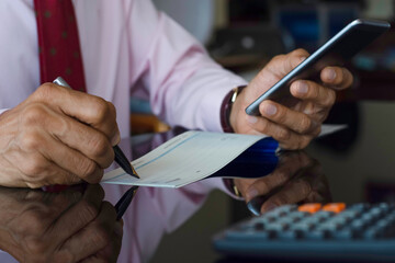 Wall Mural - Businessman hand using mobile smartphone, write and sign cheque book on the wooden table at modern home office