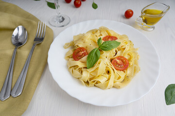 Wall Mural - Traditional italian fettucine pasta in a plate decorated with basil leaves and sliced tomatoes served together with spoon and fork on green napkin and olive oil gravy boat on white wooden background