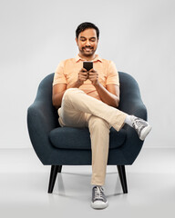 Poster - people, technology and furniture concept - happy smiling young indian man with smartphone sitting in chair over grey background