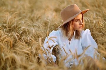 Young woman  in white linen dress and hat enjoying a sunny day in a golden wheat field. Summer, beauty, fashion, glamour, lifestyle concept.