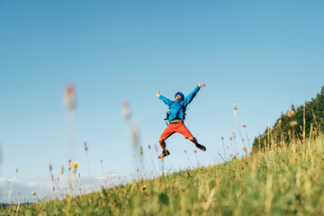 Wall Mural - backpacker traveler emotionally jumping over green grass mountain meadow with backpack with wide ope