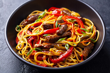 Stir fry noodles with vegetables and beef in black bowl. Slate background. Close up.