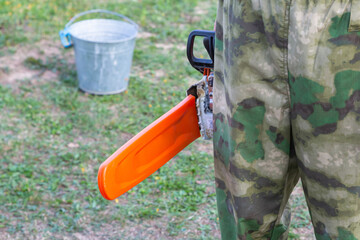 worker in camouflage uniform stands with a chainsaw, the blade in an orange protective case against the background of the earth and an iron bucket