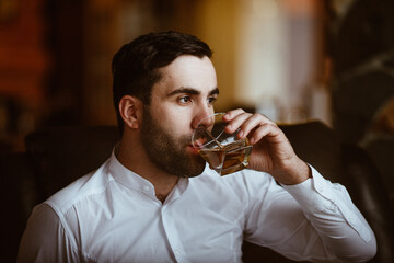 stylish charismatic man with a glass of strong alcohol in his hands sits on a chair in his large country house