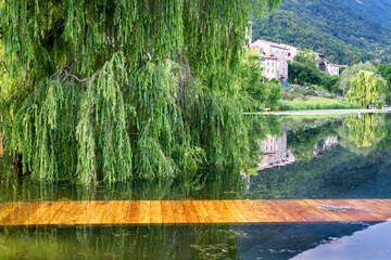 View of the village and the willow reflecting on the lake with the pier submerged in the water. View from the shore of Lago di Lago, Revine Lago, Treviso, Veneto, Italy